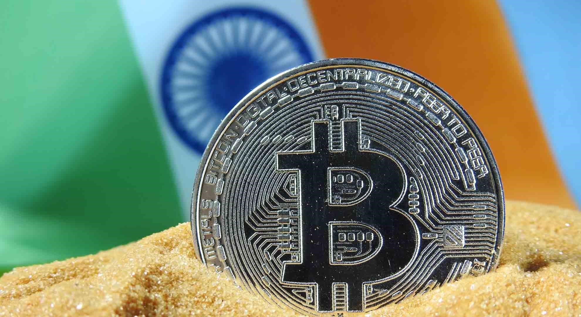 An image of a silver coin with a bitcoin logo laying in the sand, with the Indian flag in the background